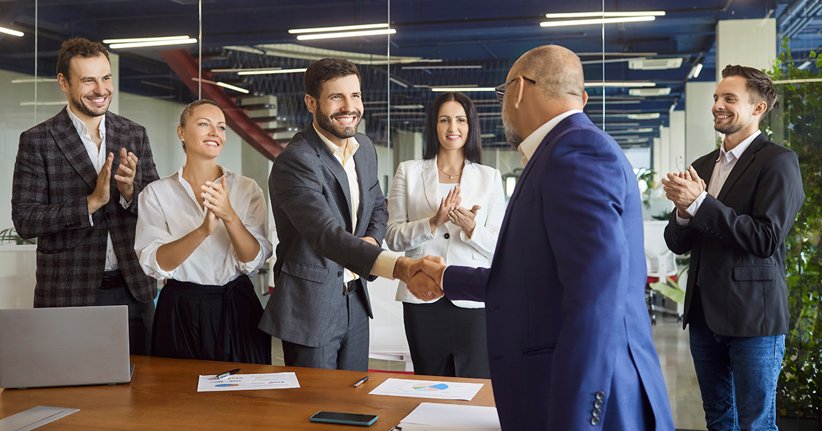 Two people performing a handshake in a meeting room with others clapping
